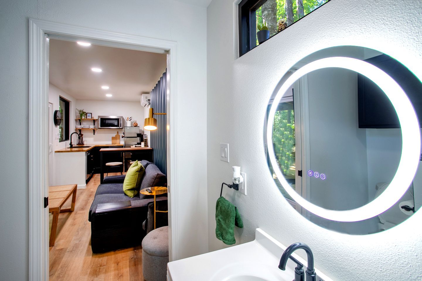 Een moderne badkamer met een ronde spiegel, een wastafel en uitzicht op een woonruimte met een bank en een eettafel.