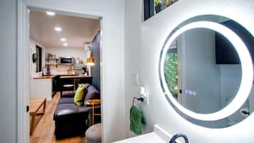 A modern bathroom with a round mirror, a sink, and a view of a living area with a sofa and a dining table.