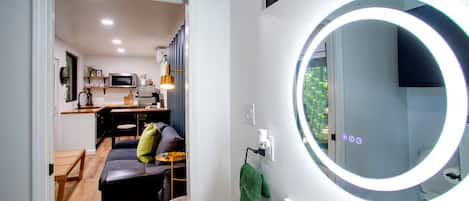 Un baño moderno con un espejo redondo, un lavamanos y vistas a una sala de estar con sofá y mesa de comedor.