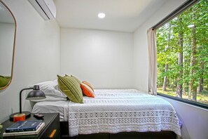 A bedroom with a bed, a bedside table with a lamp, a window with a view of trees, and a wall-mounted air conditioner.