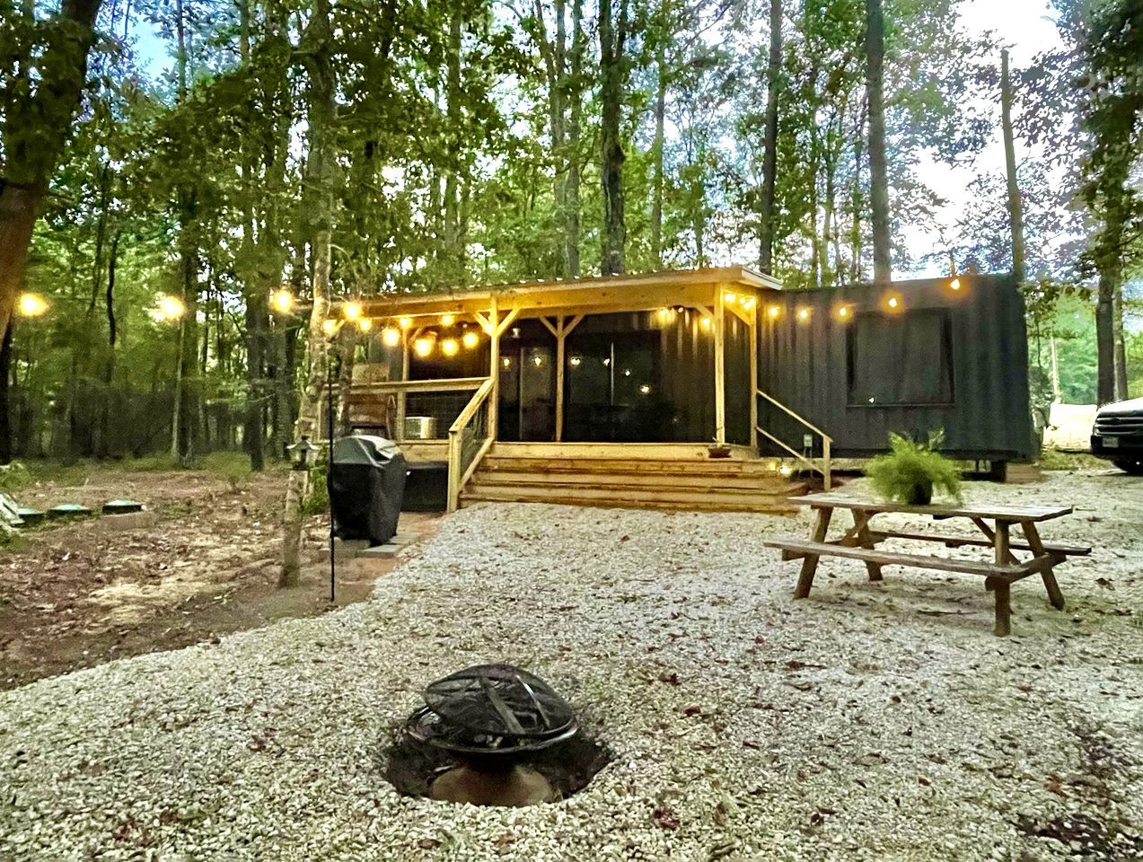 A rustic cabin with a covered porch, a picnic table, and a fire pit in a forest setting.