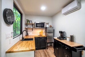 Cocina compacta con encimera de madera, armarios negros, refrigerador de acero inoxidable, microondas y un reloj en la pared.