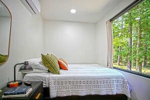 A bedroom with a bed, a bedside table with a lamp, a window with a view of trees, and a wall-mounted air conditioner. - Minimalist Luxury: Fully Equipped Modern Container, Hidden Getaway near Lake Livingston, TX (Polk County)
