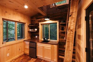 A wooden cabin kitchen with a refrigerator, sink, and windows overlooking a forest. - Magical Treehouse Stay with Creekside Sauna Experience in Zirconia, North Carolina (Zirconia)