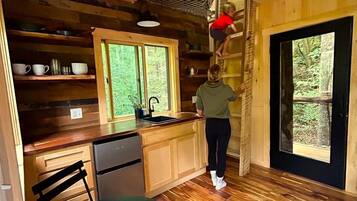 A wooden cabin interior with a kitchen, dining area, and a large window.
