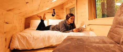 A wooden cabin with a bed, a reading chair, and a window.