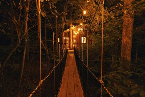 A wooden suspension bridge in a forested area with string lights. - Magical Treehouse Stay with Creekside Sauna Experience in Zirconia, North Carolina (Zirconia)