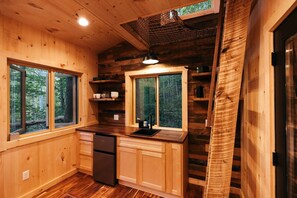 Cocina de cabaña de madera con refrigerador, lavamanos y ventanas con vistas a un bosque.