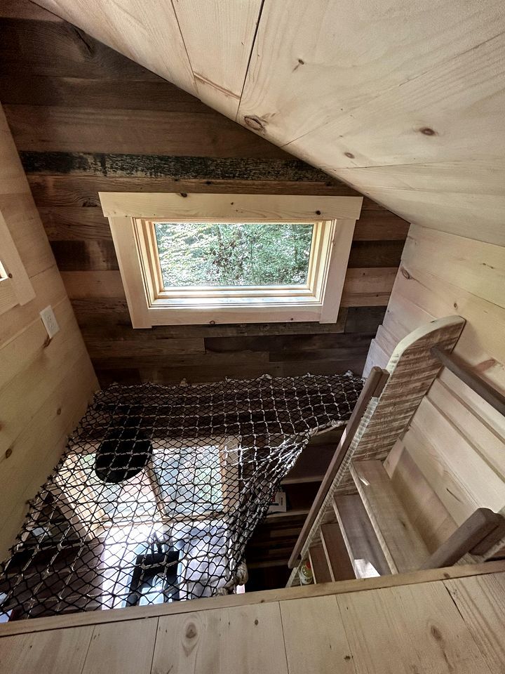 Eine Holzblockhütte mit einer Schlafgalerie, einem Fenster mit Blick ins Grüne und einem Boden aus Metallgitter.