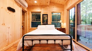 A wooden cabin bedroom with a bed, a nightstand, a lamp, and a window. - Magical Treehouse Stay with Creekside Sauna Experience in Zirconia, North Carolina (Zirconia)