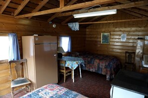 Una cabana amb un llit, una taula amb estovater, una cadira, un frigorífic i una finestra amb cortines.