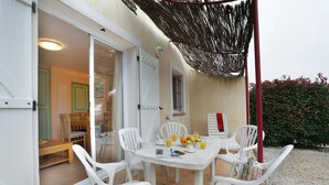 Eine Terrasse im Freien mit einem Tisch für vier Personen, umgeben von weißen Plastikstühlen, einem gewebten Sonnensegel und Blick ins Innere.