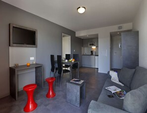 Un salon moderne avec un canapé gris, un tabouret rouge et une petite table basse. Un téléviseur à écran plat est fixé au mur, il y a un coin cuisine avec un four à micro-ondes, ainsi qu’une table à manger avec des chaises.