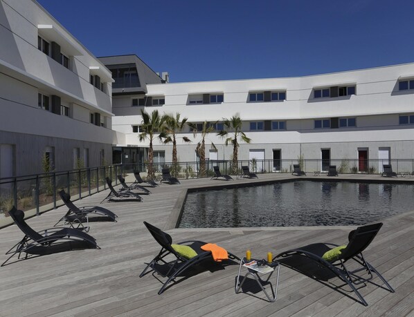 A poolside area with lounge chairs, a small table with drinks, and a building with multiple windows and balconies. - Urban Comfort Stay for 4 in Montpellier (Saint-Jean-de-Vedas)