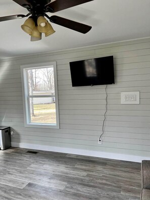 A room with a ceiling fan, a flat-screen TV mounted on the wall, a window, and a power outlet.