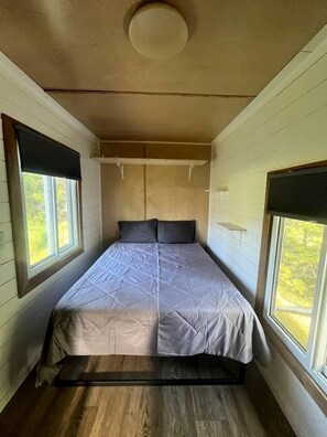 Ein kleines, gemütliches Schlafzimmer mit einem Bett, zwei Fenstern mit Jalousien, einer Deckenleuchte und Holzboden.