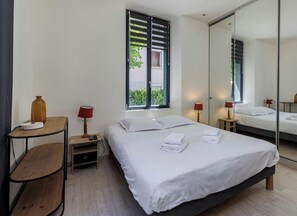 A modern bedroom with a large bed, bedside tables, a mirror, and a window with blinds. - Welcome to this modern, minimalist one-bedroom flat, perfect for a comfortable stay for two in pleasant surroundings. (Annecy)