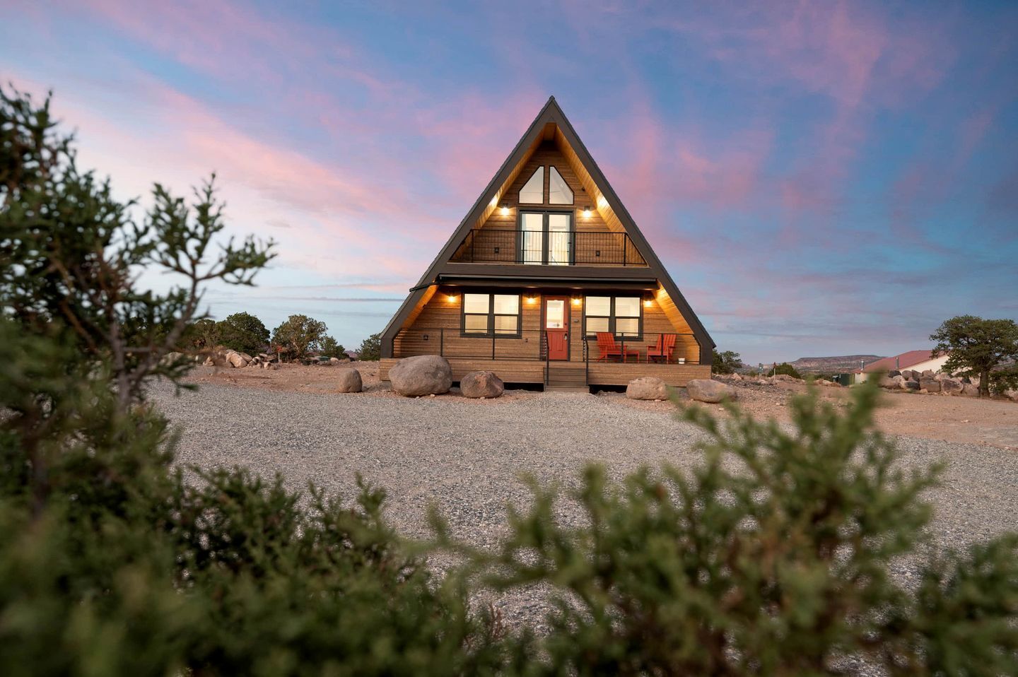Una moderna casa di legno a forma triangolare, con ampie finestre e un balcone, circondata da un vialetto sterrato e vegetazione rada.