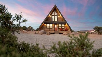 Una moderna casa di legno a forma triangolare, con ampie finestre e un balcone, circondata da un vialetto sterrato e vegetazione rada.