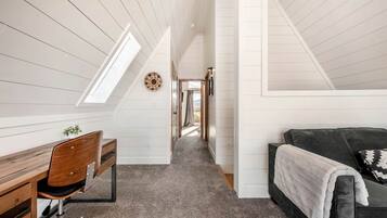 A narrow attic room with a sloped ceiling, a desk with a chair, a couch with a blanket, and a hallway leading to a door.