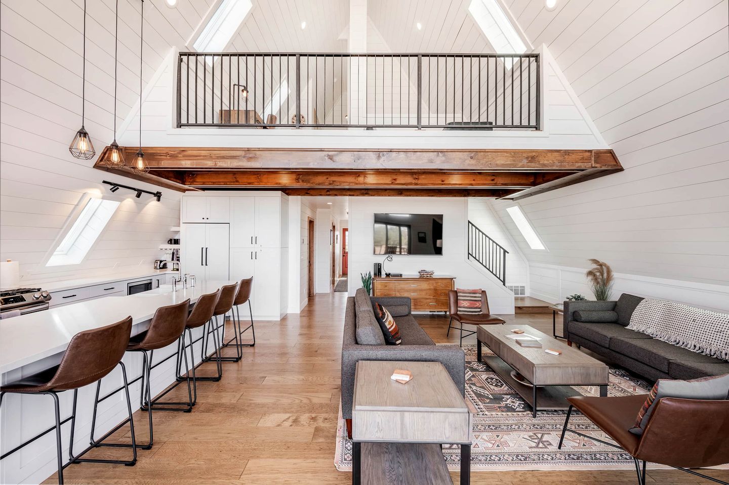 A spacious loft with a high ceiling, a kitchen with bar stools, a dining area with a wooden table, a living room with a sofa, and a staircase leading to a lofted balcony.