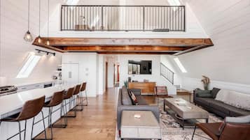 A spacious loft with a high ceiling, a kitchen with bar stools, a dining area with a wooden table, a living room with a sofa, and a staircase leading to a lofted balcony.