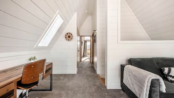 A narrow attic room with a sloped ceiling, a desk with a chair, a couch with a blanket, and a hallway leading to a door.