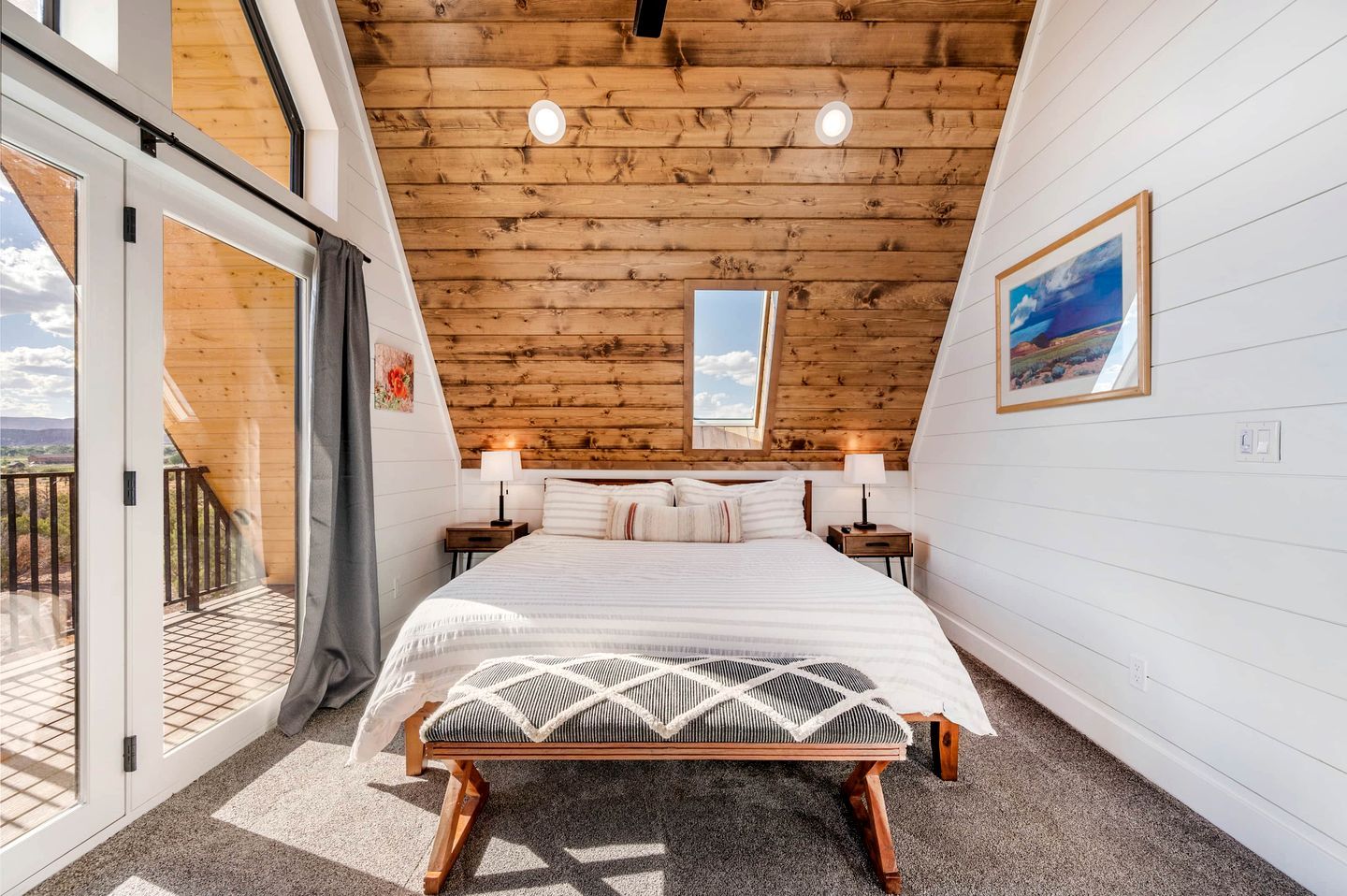 A bedroom with a large bed, two bedside tables with lamps, a wooden ceiling, and a balcony with a view.