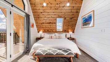 A bedroom with a large bed, two bedside tables with lamps, a wooden ceiling, and a balcony with a view.