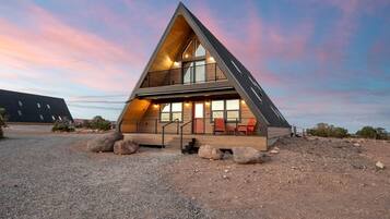 A modern, triangular-shaped cabin with a wooden exterior, a balcony, and a gravel driveway.