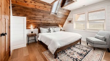 A bedroom with a sloped ceiling, a bed, a nightstand, a chair, and a window with blinds.