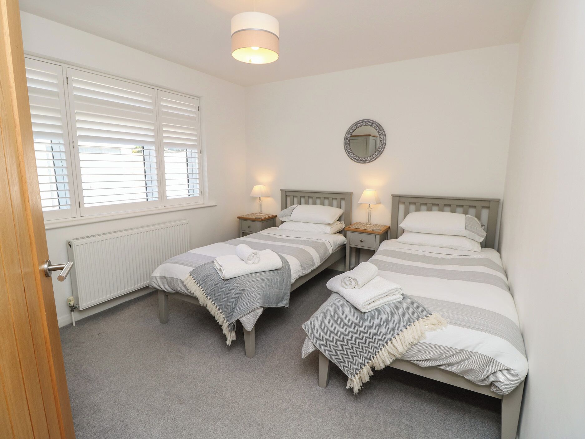 A bedroom with two single beds, a bedside table with a lamp, a mirror on the wall, and a window with blinds.