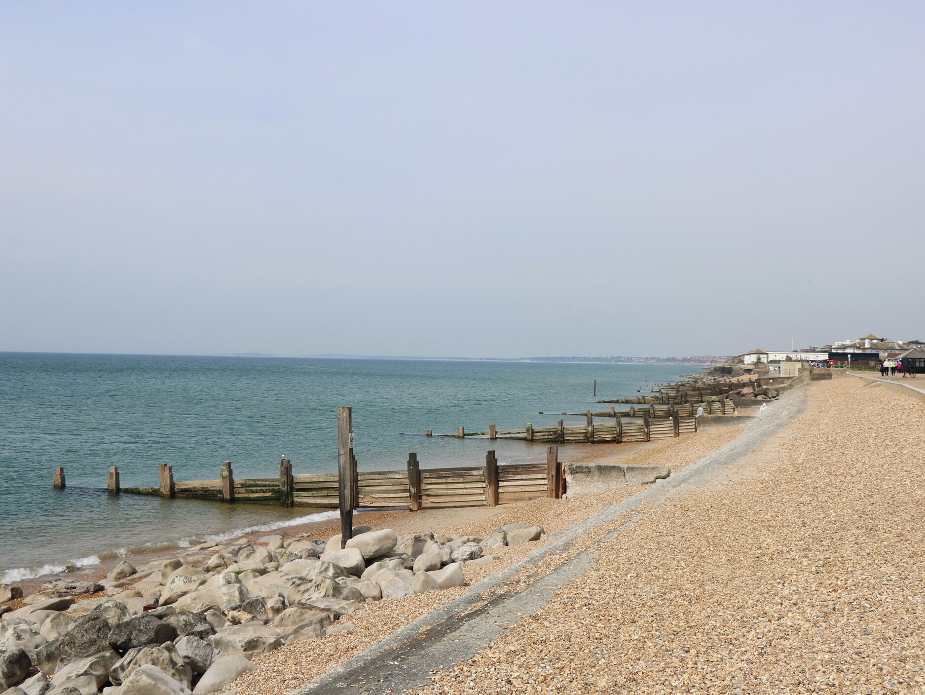 Une promenade en bord de mer avec une plage de galets, des poteaux en bois et une vue lointaine sur des bâtiments.