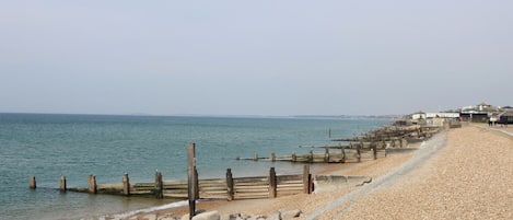 A coastal walkway with a pebble beach, wooden posts, and a distant view of buildings.