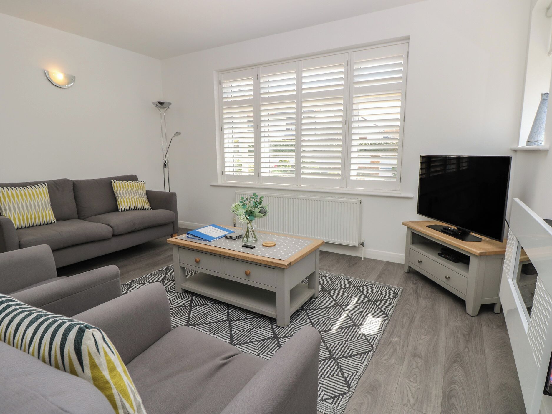 A modern living room with a grey sofa, a coffee table, a TV, and a large window with blinds.