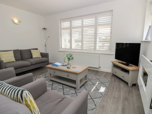 A modern living room with a grey sofa, a coffee table, a TV, and a large window with blinds. - 50 Sea Road (Lymington)
