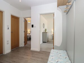 A hallway with wooden flooring, white walls, and a mirror. There is a bed with a bedside table and lamp in the background. - 50 Sea Road (Lymington)