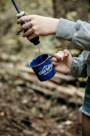 Una persona sosteniendo una taza azul con el texto 'Cold Spring Base Camp', vertiendo líquido desde una botella.