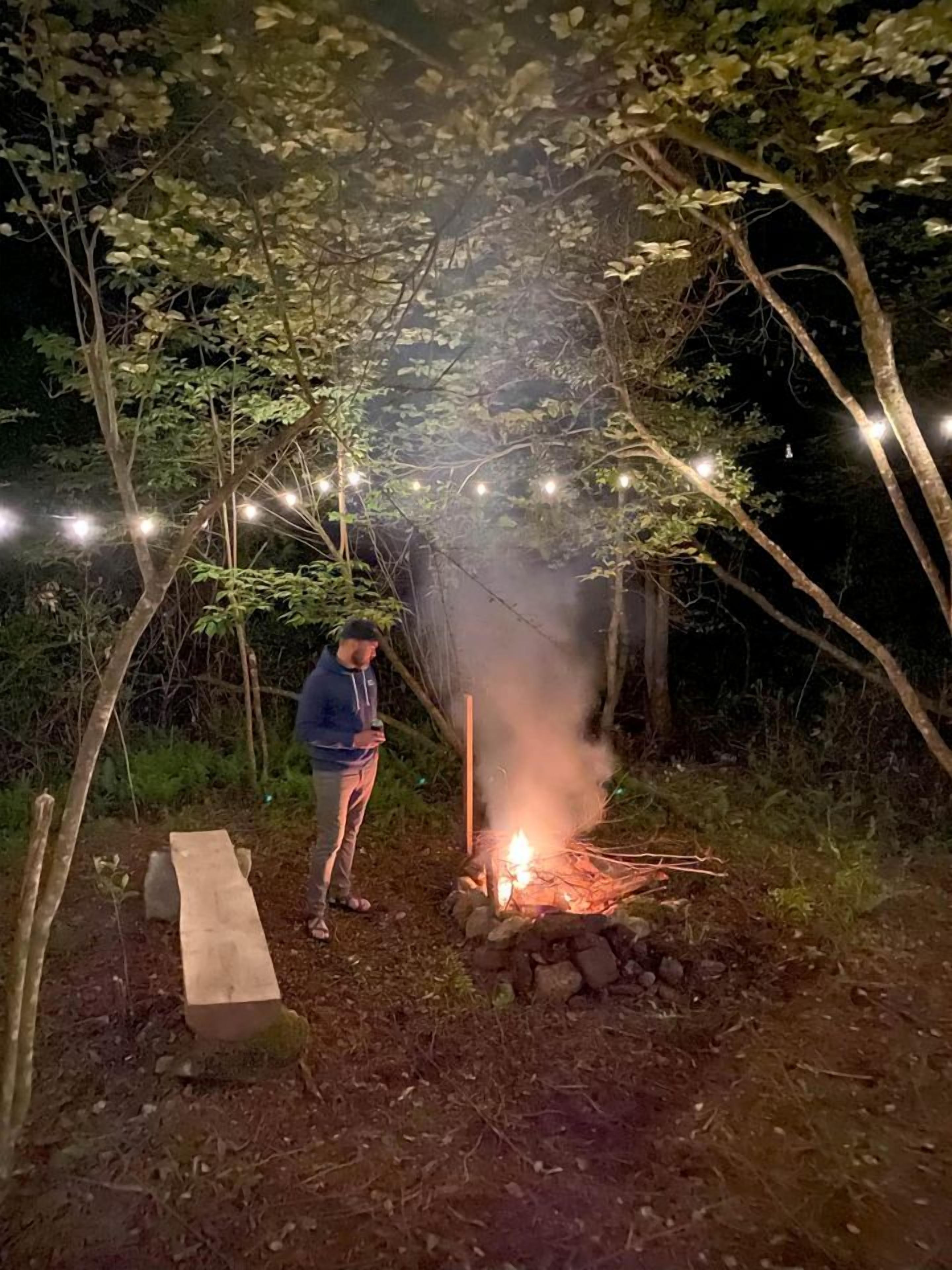 A person standing near a campfire in a wooded area with string lights overhead.