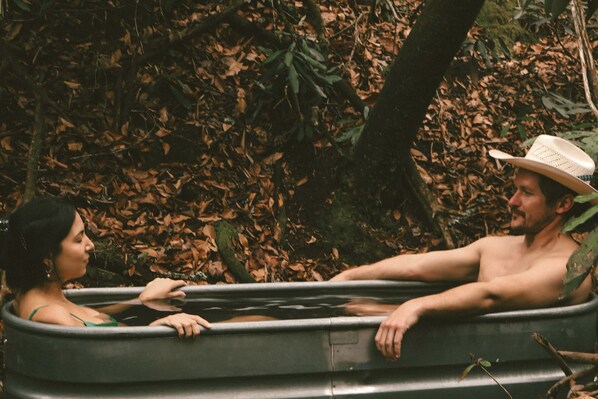 A man and a woman in a bathtub outdoors surrounded by trees.