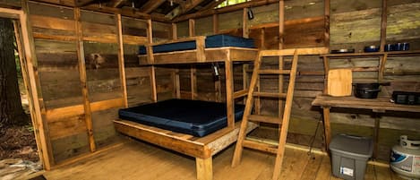 Eine rustikale Holzhütte mit Etagenbett, einer Leiter, einem kleinen Tisch mit Geschirr und einem Mülleimer.