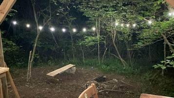 Un'area esterna con un molo di legno, un tavolo e sedie, circondata da una zona boschiva con luci decorative.