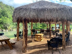 A thatched-roof pavilion with outdoor seating, a picnic table, and a view of a lake. - Cool Bell Tent #H and Cabana #12 near New Braunfels, Texas (Kingsbury)