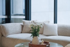 A beige sofa with white cushions, a glass coffee table with a vase of white flowers, and a wooden tray with a candle and a figurine. - Southbank Urban Haven! (Southbank)