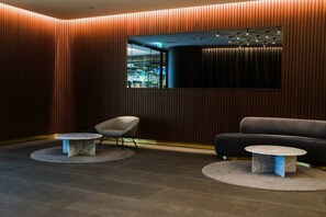 Eine moderne Lobby mit einer Wand aus Holzpaneelen, einem großen Spiegel, einer Couch, zwei runden Tischen und einem Sessel.