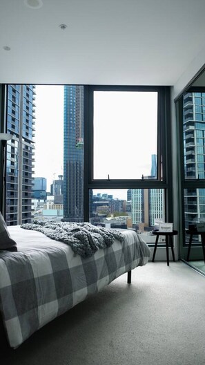 Ein Schlafzimmer mit großem Fenster, einem Bett und Blick auf hohe Gebäude.