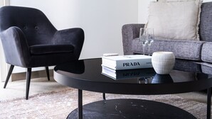 Ein modernes Wohnzimmer mit einem schwarzen Sessel, einem grauen Sofa, einem schwarzen Couchtisch mit Büchern und einer dekorativen Schale, sowie einem weißen Kissen auf dem Sofa.