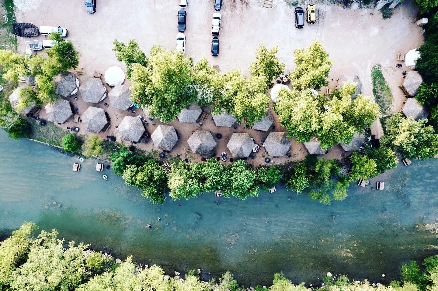 An aerial view of a riverside hotel with thatched-roof huts, a parking area, and a river.