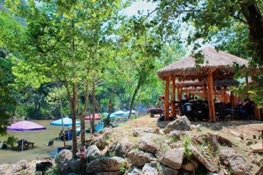 A riverside area with a thatched-roof pavilion, picnic tables, and umbrellas. - Tent #L & Cabana #17's Ultimate Family Glamping Getaway in Kingsbury, TX (Kingsbury)