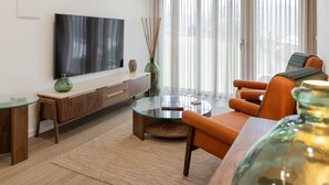 Ein modernes Wohnzimmer mit Flachbildfernseher, Glastisch, einer Couch und einem großen Fenster mit Vorhängen.